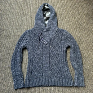 Blå/grå stickad hoodie med knappar - Säljer en sjukt snygg grå/blå stickad hoodie med ett flätat mönster och knappar framtill. Tröjan har långa ärmar och en mysig huva med snörning. Perfekt för kyliga dagar! Den har även feta elbow patches och samma mönster i luvan! Hör gärna av er vid frågor. 