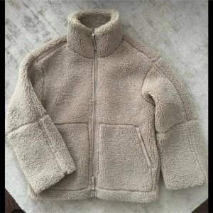 Beige teddyjacka med dragkedja - Mysig beige teddyjacka med hög krage och dragkedja framtill. Jackan har två stora fickor och är perfekt för kyligare dagar. Den har en avslappnad passform och är superbekväm att bära.