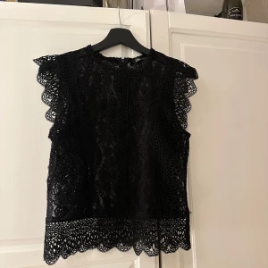 Svart spetsblus - Elegant svart spetsblus med vackra detaljer och ärmlös design. Perfekt för en stilren look med fina spetskanter runt ärmar och nederkant.