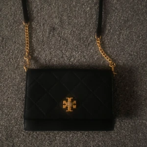 Svart quiltad axelväska från Tory Burch - Snygg svart axelväska från Tory Burch med quiltad design och guldfärgade kedjedetaljer. Väskan har en elegant logotyp framtill och en praktisk storlek för alla dina nödvändigheter. Perfekt för att ge din outfit en lyxig touch.