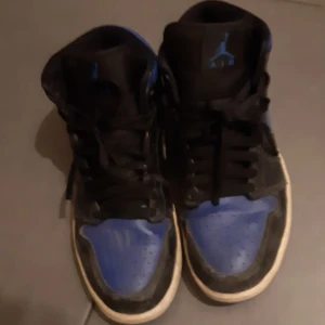 Svart och blå Nike Air Jordans - Säljer ett par klassiska Nike Air Jordans i svart och blått. Skorna har en hög design med snörning och den ikoniska Air Jordan-loggan på sidan. Perfekta för sneakerheads som älskar en tidlös stil.