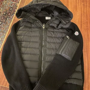 Moncler cardigan - Snygg svart jacka från Moncler med quiltad design och dragkedja. Jackan har en huva och en praktisk ficka med dragkedja på ärmen. Perfekt för kyligare dagar.