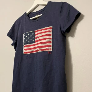 Mörkblå t-shirt med amerikansk flagga från Polo - Snygg mörkblå t-shirt från Polo med en stor amerikansk flagga på bröstet. Flaggan har en vintage-look med broderade detaljer och RL-logga. Perfekt för en avslappnad stil.