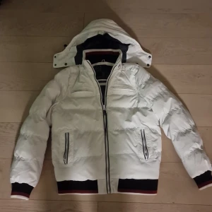 Vit pufferjacka från indicode jeans - Säljer en snygg vit pufferjacka från Indicode jeans. Jackan har en avtagbar huva och dragkedja framtill. Den är långärmad med ribbade muddar och nederkant i svart och rött. Perfekt för kyliga dagar!
