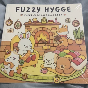 Fuzzy Hygge Super Cute Coloring Book - En charmig målarbok med mysiga hygge-teman. Omslaget visar söta djur som njuter av en varm och hemtrevlig atmosfär framför en brasa. Perfekt för avkoppling och kreativitet.
