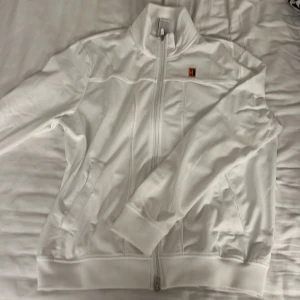 Vit jacka från Nike - Säljer en stilren vit jacka från Nike med dragkedja och hög krage. Jackan har ett färgglatt Nike-märke på bröstet och långa ärmar. Perfekt för en sportig look!