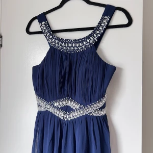 Marinblå balklänning med pärldetaljer - Elegant marinblå balklänning med vackra pärldetaljer runt halsringningen och midjan. Klänningen har en plisserad design och breda axelband, dubbelt tyg och grå ner till fötterna. Perfekt för en speciell kväll. Endast använd en gång! Storlek M, passar mig som har S. Midja: ca 70-75cm. Längd: 140cm.