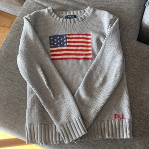 Grå stickad tröja från Ralph Lauren - Säljer en stilren grå stickad tröja från Ralph Lauren med ett amerikanskt flaggmotiv på framsidan. Tröjan har långa ärmar och ribbade kanter vid hals, ärmslut och nederkant. Perfekt för en avslappnad och trendig look.