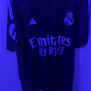 Real Madrid fotbollströja med Bellingham 5 - Snygg Real Madrid tröja från Adidas med Bellingham och nummer 5 på ryggen. Tröjan är kortärmad och har klubbens emblem på framsidan tillsammans med sponsorlogga. Perfekt för fans av laget!