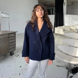 Mörkblå kappa med knappar - Snygg mörkblå kappa med dubbelknäppning och bred krage. Jackan har långa ärmar med dekorativa knappar vid ärmsluten. Perfekt för en stilren look.