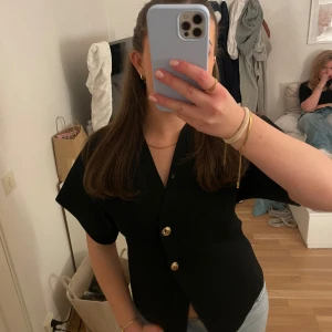 Svart v-ringad topp från Zara - Säljer en stilren svart v-ringad topp från Zara med korta ärmar och eleganta guldknappar framtill. Perfekt för en chic look.