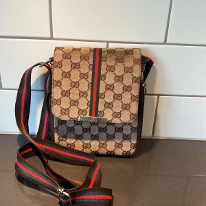 Brun axelväska med monogram - Snygg brun axelväska med klassiskt monogrammönster. Väskan har en justerbar axelrem i grönt och rött, samt en praktisk dragkedja på baksidan. Insidan är fodrad med ett mönstrat tyg och har flera fack för enkel organisering.