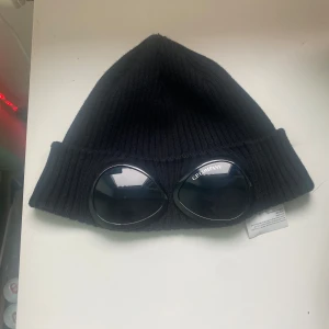 Svart mössa från C.P. Company - Snygg svart mössa från C.P. Company med unika glasögondetaljer framtill. Mössan är ribbad och tillverkad i ett mjukt material som håller dig varm. Perfekt för att ge din outfit en cool touch.