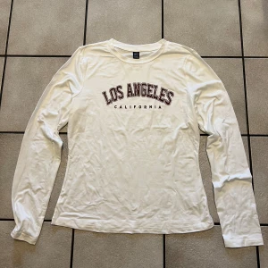 Vit långärmad tröja med tryck storlek XL - Säljer en vit långärmad tröja med texten 'Los Angeles California' tryckt på framsidan. Perfekt för en avslappnad stil. Tröjan har en rund halsringning och är gjord i ett mjukt material.