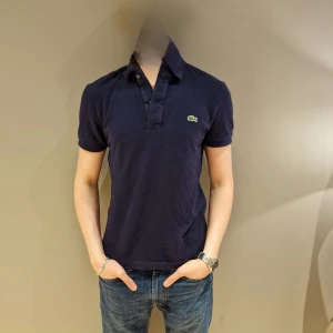 Mörkblå pikétröja från Lacoste - Snygg mörkblå pikétröja från Lacoste med klassisk krage och korta ärmar. Perfekt för en stilren och avslappnad look. Den ikoniska krokodilloggan pryder bröstet.