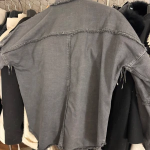 Grå jeansjacka med fransar - Snygg grå jeansjacka från Zara med coola fransdetaljer i silver längs bröstet. Jackan har knappar framtill och långa ärmar. Perfekt för att ge din outfit en unik touch!