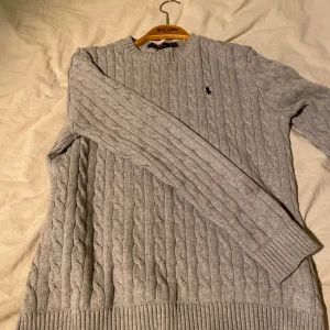 Grå stickad tröja från Ralph Lauren - Säljer en stilren grå stickad tröja från Ralph Lauren med klassiskt kabelstickat mönster. Tröjan har långa ärmar och en broderad logotyp på bröstet. Perfekt för kyligare dagar. Storlek är  M men den kan även passa dig som har S