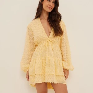 Gul klänning med volanger - Charmig gul klänning med volanger och knytning framtill. Den har en v-ringad design och långa ärmar, perfekt för vår och sommar. Den luftiga stilen ger en lekfull känsla. Passar xs och s