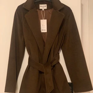 Brun kappa från NA-KD - Snygg brun kappa från NA-KD med bälte i midjan. Perfekt för höst och vinter. Klassisk design med långa ärmar och krage. Ett stilrent val för kyligare dagar. Helt ny och oanvänd.