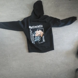 Svart hoodie med blommotiv - Säljer en svart hoodie med texten 'Optimistic' och ett blommotiv på ryggen. Framsidan har en liten textdetalj. Tröjan har långa ärmar och en praktisk ficka framtill. Perfekt för en avslappnad stil.