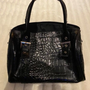 Svart krokodilpräglad handväska - Snygg svart handväska med krokodilpräglat mönster och stiliga spänndetaljer. Väskan har två handtag och en dragkedja upptill. Perfekt för att ge en elegant touch till din outfit.