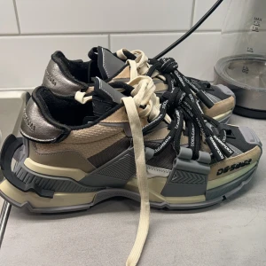 Dolce & Gabbana sneakers med futuristisk design - Snygga sneakers från Dolce & Gabbana med en unik och futuristisk design. Skorna har en kombination av beige, svart och grå färger med tydliga logotyper på snörningen. De har en robust sula och detaljerade paneler i olika material.