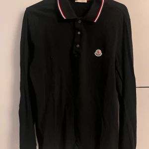 Svart långärmad pikétröja från Moncler - Snygg svart långärmad pikétröja från Moncler med klassisk krage och knappar framtill. Tröjan har en broderad logotyp på bröstet och röd-vit-blå randig detalj på kragen. Perfekt för en stilren look.