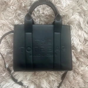 Svart axelväska från Marc Jacobs - Säljer en stilren svart axelväska från Marc Jacobs. Väskan har en präglad text 'The Tote Bag' på framsidan och är tillverkad i skinn. Den har både handtag och en justerbar axelrem för flexibel användning. Perfekt för att bära dina dagliga nödvändigheter med stil. Oanvänd helt ny
