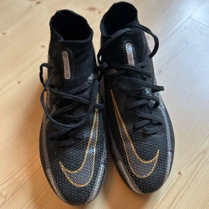 Svarta fotbollsskor från Nike - Snygga svarta fotbollsskor från Nike med gulddetaljer och snörning. Skorna har en strumpliknande passform och är designade för optimal prestanda på planen. Perfekta för fotbollsspelare som vill ha stil och funktion.
