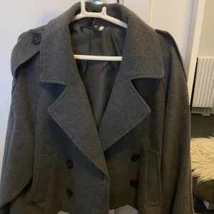 Grå kappa med knappar - Snygg grå kappa med dubbelknäppning och breda slag. Perfekt för höst och vår. Klassisk design med en modern twist, passar till många olika outfits.
