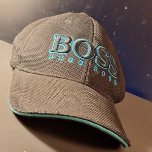 Svart blå keps från Hugo Boss - Snygg svart keps från Hugo Boss med turkos broderad logotyp framtill. Kepsen har en justerbar rem baktill med metallspänne och turkosa detaljer. Perfekt för en stilren look. Kepsen är I övrigt I mycket bra skick. Endast använd ett fåtal gånger.