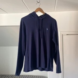 Mörkblå hoodie från Polo Ralph Lauren - Stilren mörkblå tunn hoodie från Polo med en liten broderad logga på bröstet. Tröjan har långa ärmar och en praktisk huva med dragsko. Perfekt för en avslappnad look.