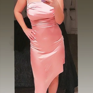 Rosa sidenklänning med smala axelband - Elegant rosa klänning i siden med smala axelband och draperad halsringning. Perfekt för en festlig kväll med sin glänsande finish och asymmetriska nederkant. Klänningen har en figurnära passform som framhäver silhuetten.