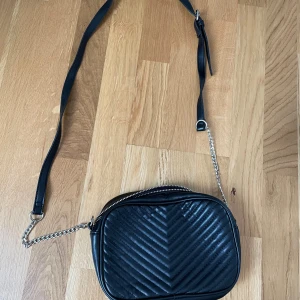 Svart väska - En svart väska med kedjedetaljer på axelremmen. Väskan är i en mönstrad läderimitation. Rymlig väska med innerfickor. Köpt utomlands. 