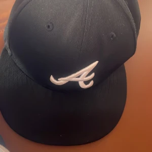 Svart keps från New Era - Säljer en svart keps från New Era med en vit broderad logga på framsidan och MLB-logga på baksidan. Kepsen är i storlek 7 (55.8 cm) och är i bra skick. Perfekt för vardagsbruk eller sportevenemang!