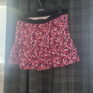 Blommig kjol från Gina Tricot - Säljer en superfin blommig kjol från Gina Tricot i storlek M. Kjolen är kort och har ett härligt blommönster i rosa och svart. Perfekt för sommarens alla fester! Den har en volangdetalj som ger den en extra söt touch. 