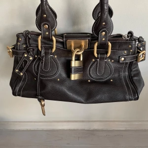 Brun väska - En cool brun väska med guldiga detaljer och ett guldigt lås. Det hänger också en nyckel på som låser upp låset om man inte vill ha det på. Inga stora skador eller stora tecken på att den är använd. Använt kanske 5-7 gg.Pris kan diskuteras 