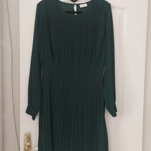 Mörkgrön klänning med långa ärmar - Säljer en superfin mörkgrön klänning med långa ärmar från VILA.  Klänningen har en elegant plisserad design och en knappdetalj vid ärmsluten. Den har en liten öppning med knapp i nacken för en extra touch. dragkedja vid sidan. Perfekt för sommar, höst och vinter, och kan användas både till vardags och fest. Passar bra till både stövlar och klackar som till en sandal eller sneakers.  den är i storlek L men klär fint en M eller S