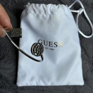 Silverfärgat armband från Guess - Säljer ett elegant silverfärgat armband från Guess. Armbandet har en öppen design med två runda detaljer med ett utskuret mönster. Perfekt för att ge en stilren touch till din outfit. Kommer med originalpåse och certifikat💕