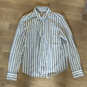 Randig skjorta från Levi's - Säljer en snygg randig skjorta från Levi's i storlek XS. Skjortan är vit med blå ränder och har långa ärmar. Den är gjord av bomull och har knappar hela vägen ner. Perfekt för både vardag och fest!