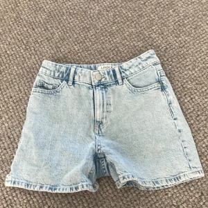 Jeansshorts Lindex  - Säljer ett par ljusblå jeansshorts från Lindex i storlek EUR 146 / UK 10-11Y. Shortsen har en klassisk design med fem fickor och knappstängning fram. Perfekta för sommaren och kan matchas med nästan vad som helst. De är i bra skick och redo för en ny ägare!