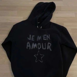 Svart hoodie med text - Säljer en svart hoodie med texten 'Je m'en amour' och en stjärna på framsidan. Den är supermjuk och perfekt för kyliga dagar. Hoodien har en stor luva och långa ärmar. Perfekt för en avslappnad och bekväm stil!