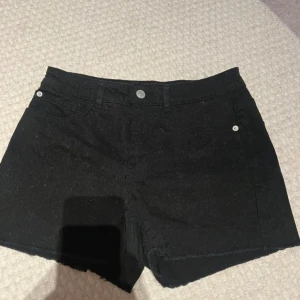 Svarta jeansshorts - Säljer ett par svarta jeansshorts i nyskick. De är högmidjade och har en knapp och dragkedja framtill. Shortsen har en normal passform och är perfekta för sommaren. De har också två fickor fram och två bak.