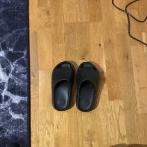 Svarta sandaler från Adidas - Säljer ett par svarta sandaler från yeezy.De är i bra skick och har en enkel och stilren design. Perfekta för sommar och bad. Sandalerna har en räfflad sula för bättre grepp och komfort.