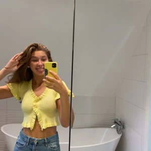 Gul ribbad topp med volangdetaljer - Säljer en superfin gul ribbad topp med volangdetaljer. Toppen har korta ärmar och en v-ringad design med knappar framtill. Perfekt för sommaren och kan stylas både till vardags och fest. Toppen är i nyskick och väldigt bekväm att bära. Lite liten i storleken🌼