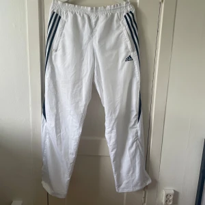 Vita träningsbyxor från Adidas - Ett par vita träningsbyxor från Adidas i storlek M. Byxorna har en elastisk midja och är gjorda av ett lätt och andningsbart material. Svart ränder på sidorna.