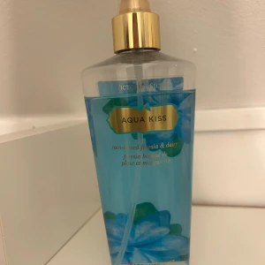 Aqua Kiss Fragrance Mist från Victoria's Secret - Säljer en fräsch och blommig Aqua Kiss Fragrance Mist från Victoria's Secret. Den har en doft av regn-kysst fresia och prästkrage. Flaskan är genomskinlig med blått innehåll och har en guldfärgad spraypump. Formulan är berikad med aloe vera och lugnande kamomill. Perfekt för en uppfriskande doft. Flaskan är 250 ml.