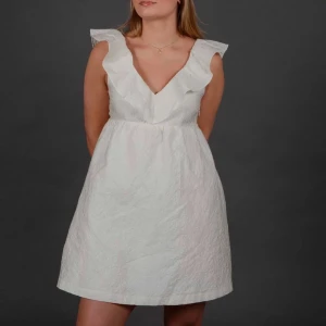 Vit klänning med volangdetaljer - Säljer en söt vit klänning med volangdetaljer och djup v-ringning. Klänningen har en kort längd och är ärmlös, perfekt för sommarens festligheter. Materialet ger en lätt och luftig känsla.