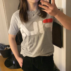 Grå t-shirt från Fila - Säljer en grå t-shirt från Fila med stort logotryck i vitt och rött på framsidan. T-shirten har en klassisk rund hals och korta ärmar. Den är knappt använd!🌟