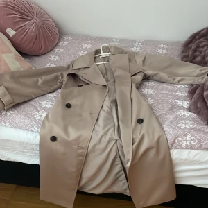 Beige trenchcoat från H&M - Snygg beige trenchcoat från H&M med klassisk design. Jackan har knappar framtill och ett bälte i midjan för en justerbar passform. Har använt ett få tals gånger inga difekter, passet till s och m